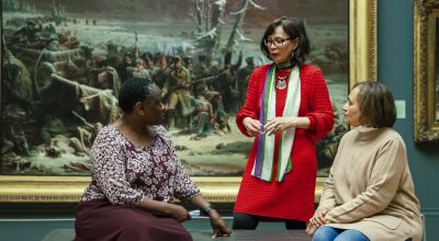 3 women in a gallery space, two are sat down on a bench and the woman in the middle is stood behind. In the background is a large framed painting