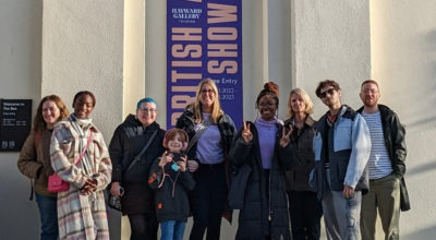 Group of people standing in front of a white wall. At the centre a purple banner with yellow writing reads: British Art Show 9