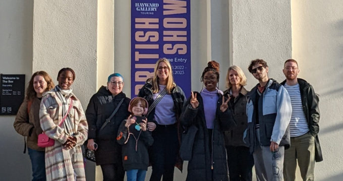 Group of people standing in front of a white wall. At the centre a purple banner with yellow writing reads: British Art Show 9