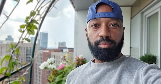 his image shows a close-up portrait of a man with a beard and baseball cap. He appears to be standing on a balcony or terrace, with a cityscape visible in the background.