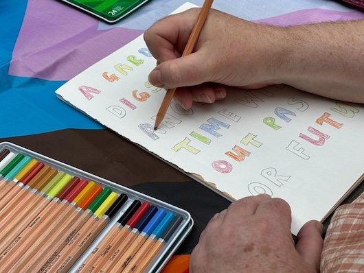 This image shows a person's hands writing on a piece of paper using coloured pencils. The paper has indistinct words written on it, along with other colourful text and designs.