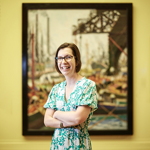 A woman with a short brown bob and glasses wearing a green floral dress, she smiles at the camera with her arms folded in front of a large painting with a yellow background