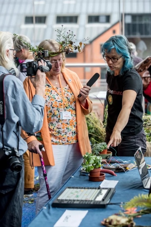 This image shows a group of people gathered around a table or display, with one person holding a microphone and interviewing another person. There are various potted plants, flowers, and gardening tools visible, suggesting this may be a horticultural or gardening event or festival. The background includes the facade of a building, indicating this is taking place outdoors in a public space.