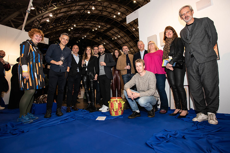 Group of people in the background that pose in a semiarch in front of a bicolored vase. The artist is sitting next to it.