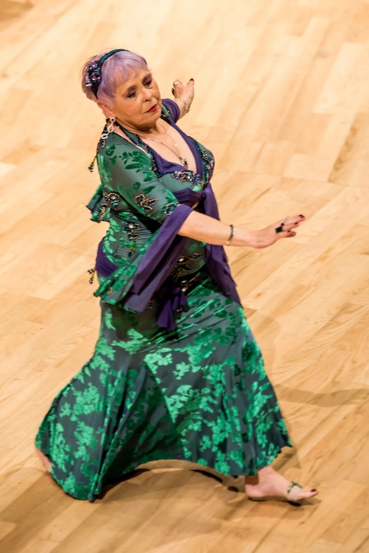 This image shows an older woman dressed in a colourful, ornate costume, presumably a traditional or cultural dress. She appears to be in the middle of a dance or performance, with a focused, expressive expression on her face. The background suggests this is taking place on a stage or in a performance venue.