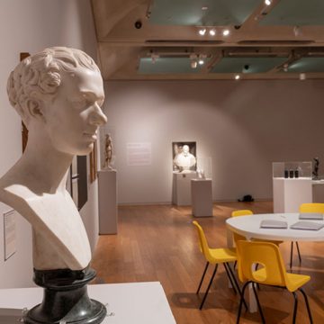 Bust of a male head on left hand side of image, in exhibition/ research room, a table is set with books and chairs placed around it