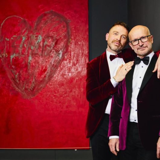 Male couple in matching velvet suits stood in front of Derek Jarman, Protest