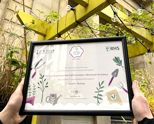 Two hands holding a sign highlights that the RHS and North West in Bloom are presenting an award as a recognition for
