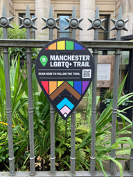 a pointed sign with the colors of the LGBTQ+ flag and the writing Manchester LGBTQ+ trail. The sign is attached to the railing of the gallery. in the background foliage and part of the facade